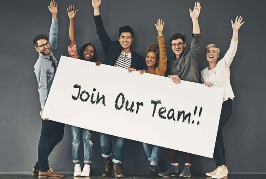 A team of young employees holding a join our team sign with their hands raised.